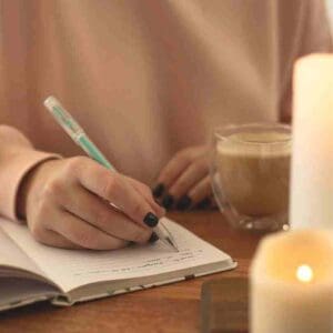 Hand writing in a notebook with a pen, coffee cup, and lit candle on wooden table.